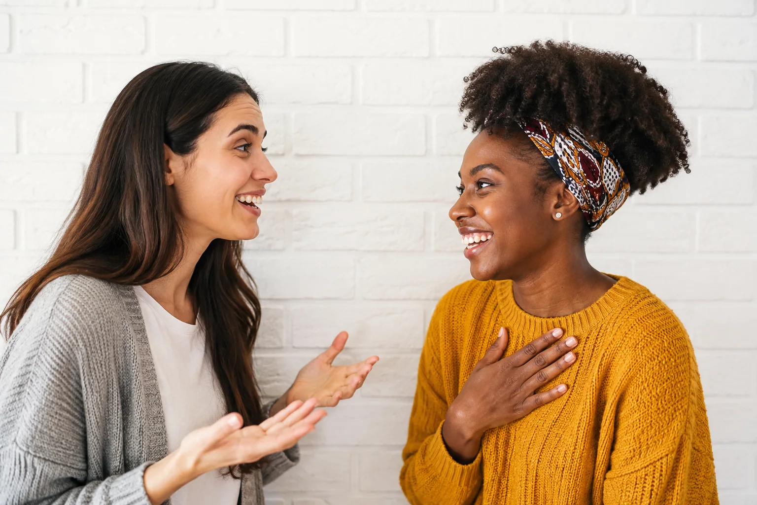 Duas amigas conversando e sorrindo diante de uma parede clara.
